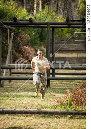 Sprinting male soldier wearing camouflage pants through grassy field toward wooden obstacle rails 129584901