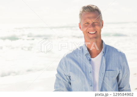 Man smiling while standing on sandy beach under sunlight wearing blue striped shirt, copy space 129584902