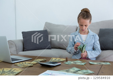 Female child counting currency bills using calculator near laptop at living room table, copy space Female child counting currency bills using calculator near laptop at living room table, copy space 129584910