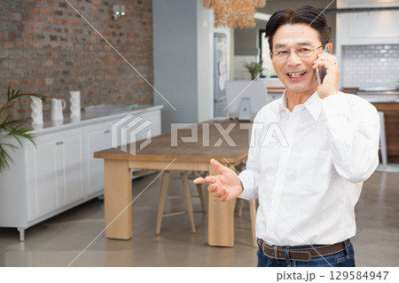 Senior Asian man standing near wooden dining table in modern kitchen holding smartphone, copy space 129584947