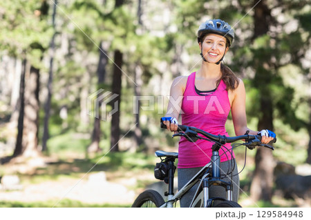 White woman cyclist posing beside mountain bike in forest wearing pink top and helmet, copy space White woman cyclist posing beside mountain bike in forest wearing pink top and helmet, copy space 129584948