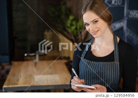 Smiling female waitress by wooden table in cafe noting orders on notepad by chalkboard, copy space 129584956