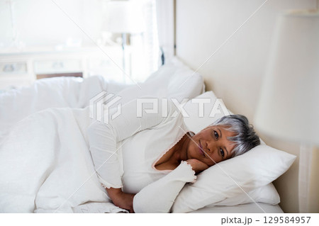 Senior woman reclining on white bed in bright bedroom, resting head on pillow near bedside lamp Senior woman reclining on white bed in bright bedroom, resting head on pillow near bedside lamp 129584957