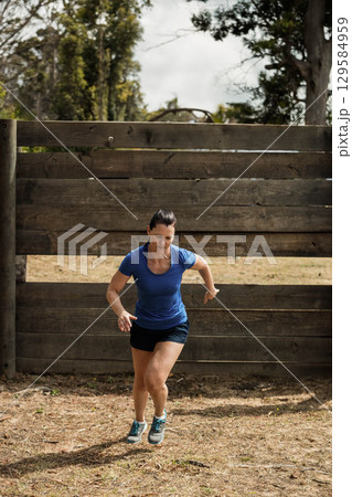 Woman vaulting wooden wall obstacle on dirt course wearing blue shirt black shorts running shoes 129584959