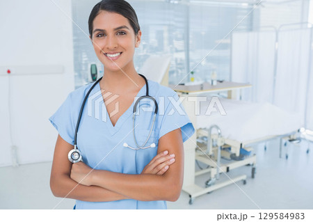 Asian woman standing with arms crossed in clinic wearing scrubs and stethoscope beside hospital bed 129584983