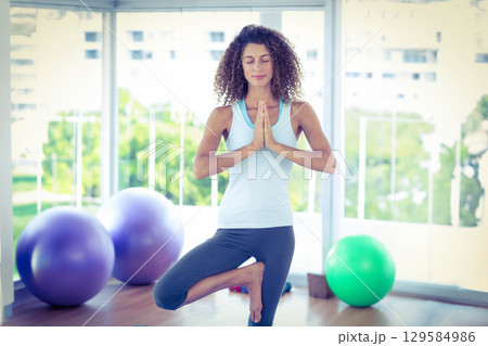 Woman practicing yoga tree pose on wooden floor by windows with purple and green exercise balls Woman practicing yoga tree pose on wooden floor by windows with purple and green exercise balls 129584986