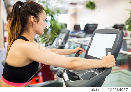 Female fitness enthusiast gripping elliptical handles monitoring digital console at gym, copy space Female fitness enthusiast gripping elliptical handles monitoring digital console at gym, copy space 129584987