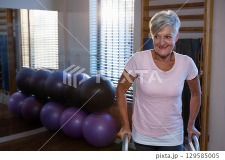 Senior woman balancing on parallel bars at gym with stability balls, mirror, blinds, copy space 129585005