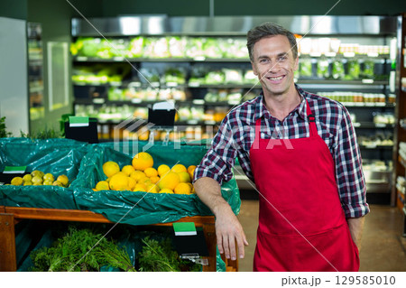 Man wearing red apron, plaid shirt leaning on bins at supermarket with oranges, lemons, copy space 129585010