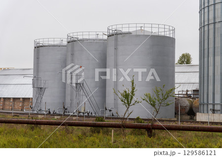 Tall grey silos/tanks in grassy field. Railway tracks, pipeline in front. Overcast 129586121