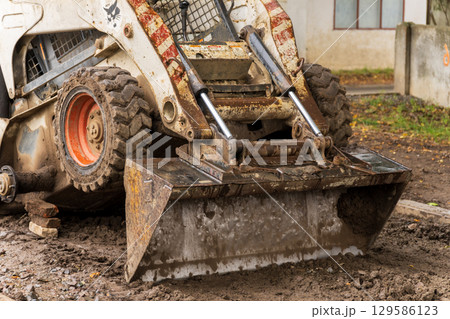 A white compact loader with red and white stripes is parked on a muddy construction site. The loader's metal bucket is covered in dirt and grime 129586123