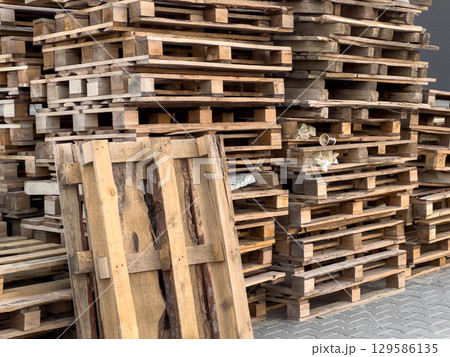 A large pile of used wooden pallets stacked on top of each other sits outdoors near a warehouse during daylight hours 129586135