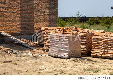 Red bricks are stacked on the ground near a brick house under construction. One brick stack is wrapped in plastic Red bricks are stacked on the ground near a brick house under construction. One brick stack is wrapped in plastic 129586154