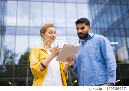 Two professionals review information on a tablet in front of a modern glass building, discussing a project. 129586457