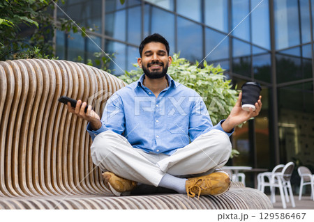 A man in a blue shirt meditates on a wooden bench, holding a coffee cup and a phone in an outdoor setting. A man in a blue shirt meditates on a wooden bench, holding a coffee cup and a phone in an outdoor setting. 129586467