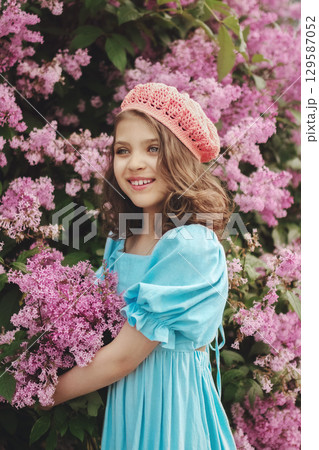Smiling girl holding flowers in blue dress and pink beret crochet against backdrop of blooming lilacs 129587052