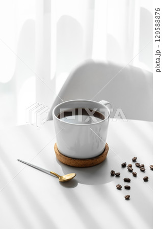 Freshly brewed espresso coffee in a cup on a white table near window with spoon, chair, coffee beans Freshly brewed espresso coffee in a cup on a white table near window with spoon, chair, coffee beans 129588876