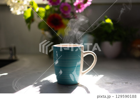 Warm Cup of Coffee on a Sunny Kitchen Counter With Flowers in the Background 129590188