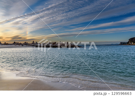 Scenic coastal view with calm sea, sandy beach, and rock formations under a vibrant sky at sunset 129596663