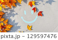 Autumn leaves in warm tones frame chalk smiley, autumn shadows stretch softly across surface, autumn mood glows through playful leaf placement and cheerful seasonal message, copy space 129597476