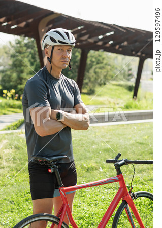 Cyclist standing wearing gray t-shirt and relax at urban city park. Athlete taking a break. Outdoor activity day off. Summer season for travel. 129597496