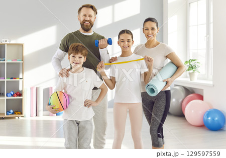 Sporty family with kids standing in gym with sports equipment in hands and looking at camera Sporty family with kids standing in gym with sports equipment in hands and looking at camera 129597559