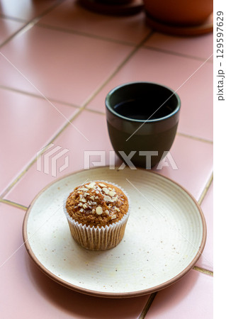 cup of coffee and muffin sit on a white plate on pink tiled table. muffin is sprinkled with nuts. 129597629