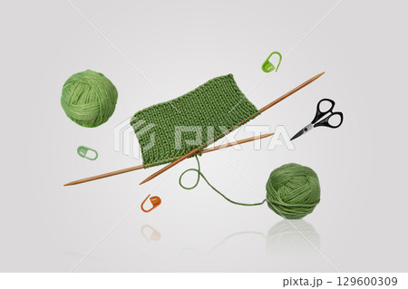 Tools for knitting warm clothes: scissors, green ball of thread, stoppers, and knitting needles with a knitted green piece of fabric flying in the air on a gray background. Hobby home crafts. 129600309