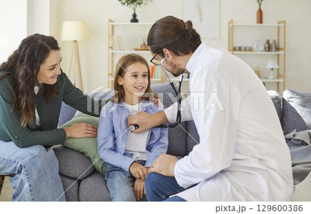 Friendly male doctor using stethoscope to check heartbeat of smiling child during home health visit. 129600386