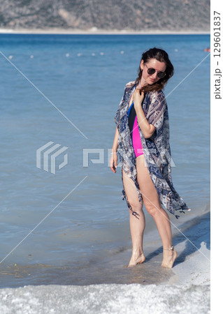 A woman dressed in a swimsuit and cover-up strolls by the waters edge, enjoying the refreshing sea breeze and warm sunshine on a beautiful day. A woman dressed in a swimsuit and cover-up strolls by the waters edge, enjoying the refreshing sea breeze and warm sunshine on a beautiful day. 129601837