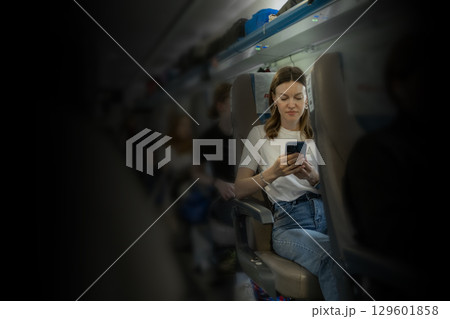 A young woman is focused on her smartphone while seated comfortably on an airplane, surrounded by other passengers during the flight. A young woman is focused on her smartphone while seated comfortably on an airplane, surrounded by other passengers during the flight. 129601858