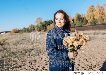 Female portrait. Beautiful young girl with long dark hair walking with bouquet of flowers along beach on sunny autumn day. High quality photo 129602794