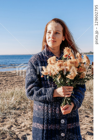 Female portrait. Beautiful young girl with long dark hair walking with bouquet of flowers along beach on sunny autumn day. Vertical photo. High quality photo 129602795