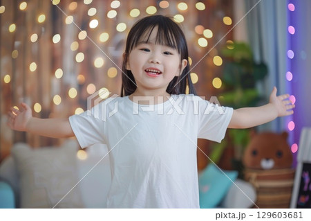 Happy Child in White T-Shirt with Open Arms in Bright Room with String Lights 129603681