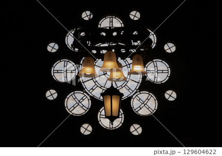 a distinctive hanging light fixture silhouetted against an ornate rose window and strong dark background. a distinctive hanging light fixture silhouetted against an ornate rose window and strong dark background. 129604622