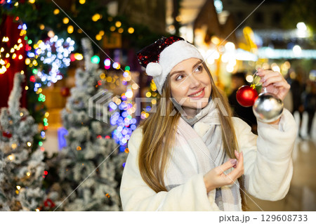 Girl in red Santa hat and with Christmas tree toys in her hands smiles dreamily Girl in red Santa hat and with Christmas tree toys in her hands smiles dreamily 129608733