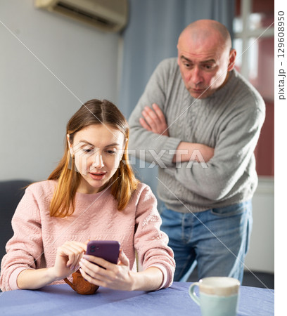 Angry husband observing his wife sitting and using phone Angry husband observing his wife sitting and using phone 129608950