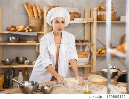 Smiling relaxed young sexy girl professional baker topless in white uniform standing at work table and kneading dough during working day in bakery 129609132