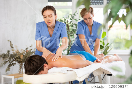 Two professional masseuses performing back massage for male patient 129609374