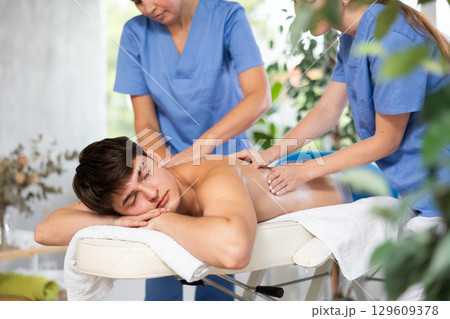 Two unrecognizable female masseuses perform stroking and rubbing of muscles of back to guy client 129609378