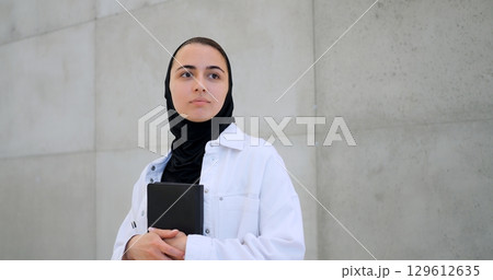 Muslim woman wearing hijab, white shirt, holding book close, gazing pensively near concrete wall, embodying contemplative moment 129612635