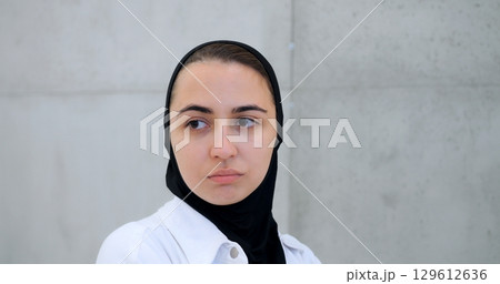 Muslim woman in hijab displaying varied facial expressions from contemplative to intense, standing against minimalist concrete wall backdrop Muslim woman in hijab displaying varied facial expressions from contemplative to intense, standing against minimalist concrete wall backdrop 129612636