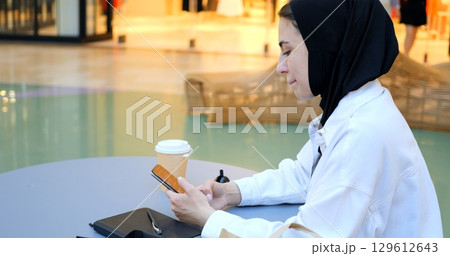 Young entrepreneur wearing hijab using smartphone, enjoying coffee break in modern office building, demonstrating focus and productivity in dynamic work environment. Freedom for Muslim women 129612643