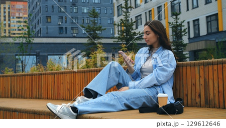 Comfortable woman relaxing on wooden bench, browsing smartphone with takeaway coffee cup nearby, enjoying peaceful moment during urban day 129612646