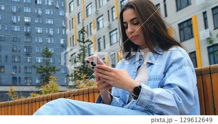 Young woman wearing casual denim jacket sitting on urban bench, browsing internet and engaging with mobile app while enjoying peaceful moment in modern city environment 129612649
