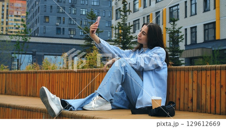 Young woman in casual attire sitting on urban bench, using smartphone for selfie, messaging, social media while enjoying coffee break amid city surroundings Young woman in casual attire sitting on urban bench, using smartphone for selfie, messaging, social media while enjoying coffee break amid city surroundings 129612669