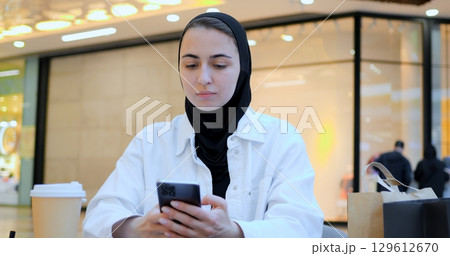 Young entrepreneur wearing hijab reviews notes and uses tablet in bustling shopping center food court, showcasing modern work practices and cultural diversity. Muslim diligent student, flexible 129612670