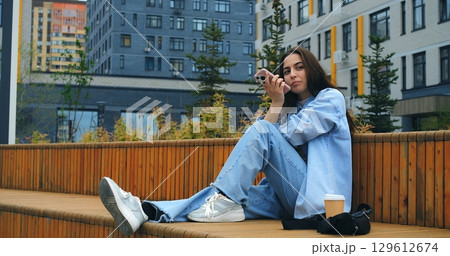Young woman, wearing casual attire, sits on a wooden bench, savoring a takeaway coffee while engaging with her smartphone against a backdrop of modern buildings Young woman, wearing casual attire, sits on a wooden bench, savoring a takeaway coffee while engaging with her smartphone against a backdrop of modern buildings 129612674