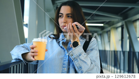 Businesswoman holding a paper cup of coffee and using her smartphone, sending voice messages and text messages and walking on a bridge in a modern city 129612678