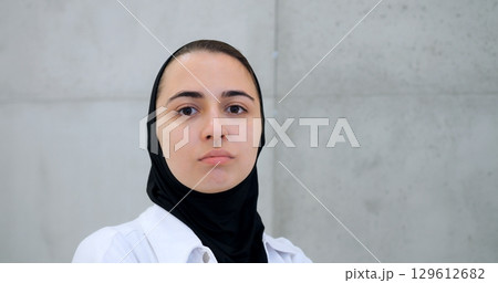 Professional muslim female healthcare worker wearing hijab and white coat, standing against concrete wall, displaying confident facial expressions while facing camera Professional muslim female healthcare worker wearing hijab and white coat, standing against concrete wall, displaying confident facial expressions while facing camera 129612682